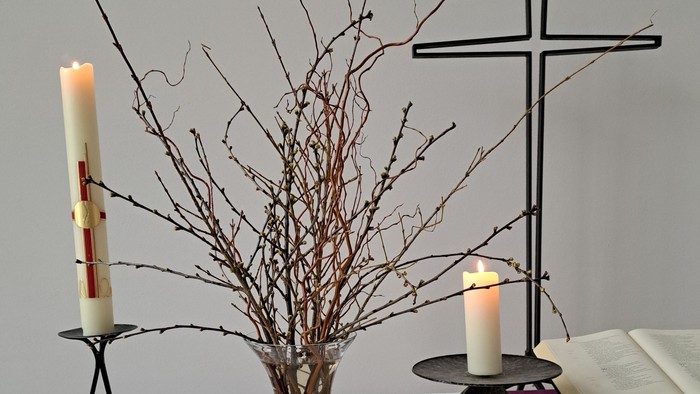 Eine ruhige Altar-Anordnung mit Kerzen, einem Kreuz, getrockneten Zweigen und einem offenen Buch auf einem bedeckten Tisch.