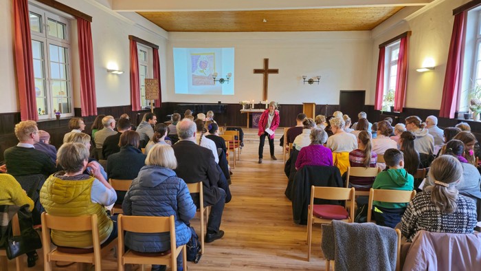 Eine Gruppe von Menschen sitzt in einem Raum mit Holzboden und hört einem Redner zu, der vor einem Bildschirm steht.