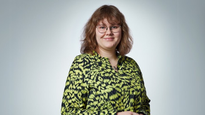 En kvinde står mod en gul væg i en grøn og sort fløjtefarvet jakke.