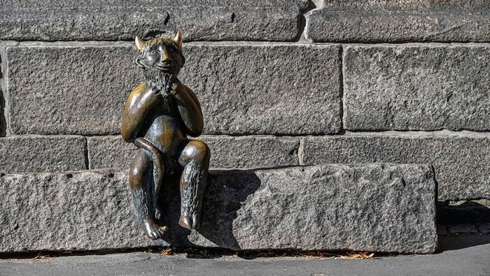 Eine verspielte Bronzestatue eines ziegenartigen Wesens, das auf einer Steinmauer sitzt.