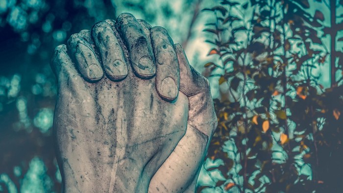 Eine Steinstatue einer Hand im Gebet, umgeben von grünen Blättern.