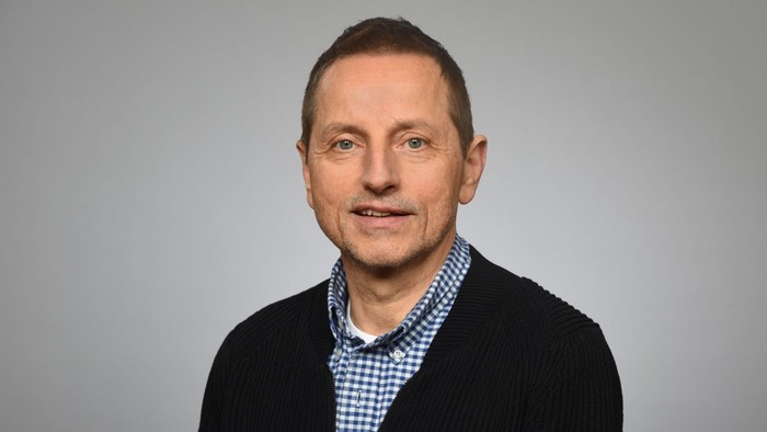 Ein Mann mit dunklem Haar und Bart, der ein schwarzes Jackett und ein blau-weiß kariertes Hemd trägt.