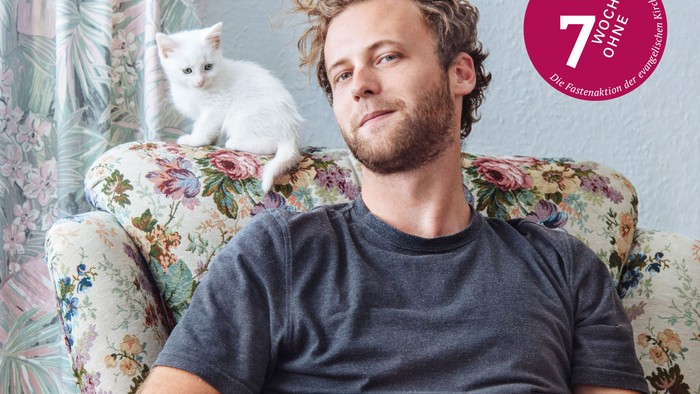 Ein Mann auf einem geblümten Sessel sitzend, auf der Rücklehne ein weißes Kätzchen