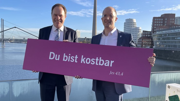 Zwei Männer halten ein Schild mit der Aufschrift „Du bist kostbar“ vor einer Stadtkulisse mit einem Turm im Hintergrund.