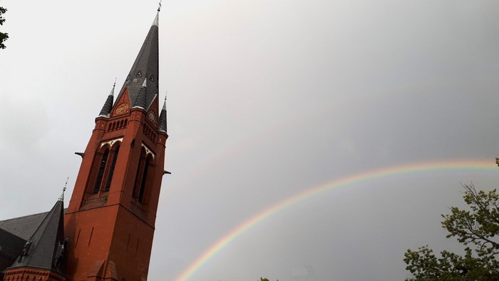 Ein roter Backsteinkirchturm ragt vor einem grauen Himmel empor, während sich daneben ein leuchtender Regenbogen über den Bäumen spannt.