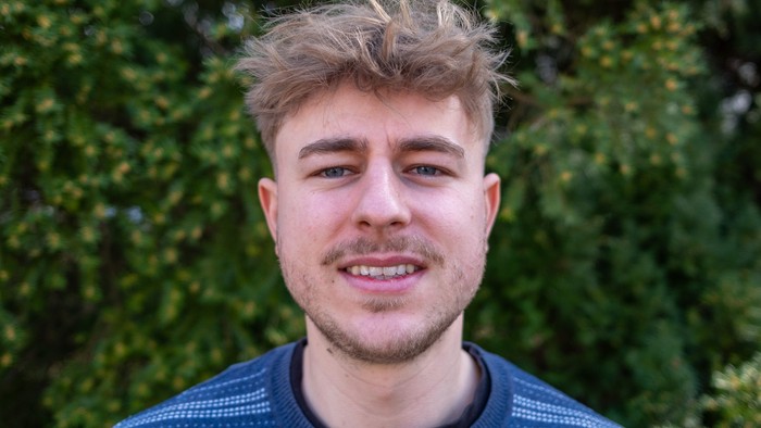 Mann, Ende 20, wuschelige blonde kurze Haare, Blick nach vorne, blauer Pullover mit weißen Querstreifen, grüne Hecke im Hintergrund