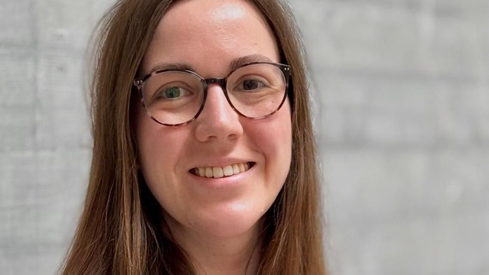 Eine lächelnde Person mit Brille, die einen rot-rosa gestreiften Pullover trägt, vor einem neutralen Hintergrund.