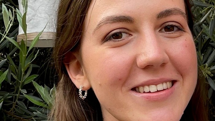 Lächelnde Frau mit welligem Haar in einer Bluse mit Blumenmuster vor üppigem grünem Blattwerk-Hintergrund.