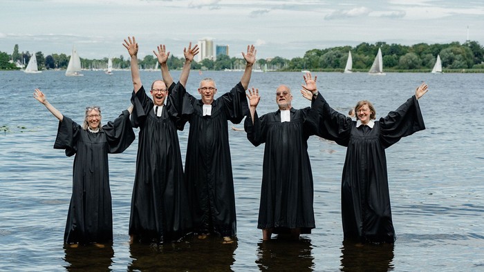 Fünf Pfarrpersonen im schwarzen Talar stehen im Wasser der Havel und strecken die Arme gen Himmel.