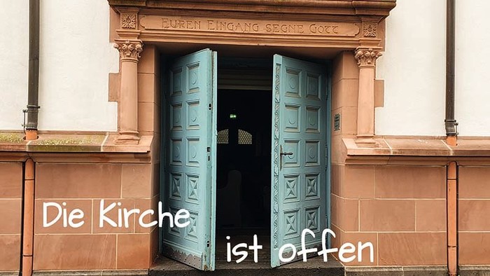 Kirchentüren mit dem Schild *„Die Kirche ist offen“* laden Besucher in den Eingang eines historischen Gebäudes ein.