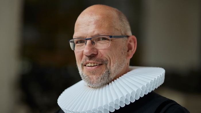 Mann mit Glatze und weißem Kragen, trägt Brille und lächelt