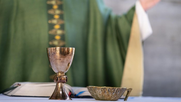 Ein Priester in grünen Messgewändern hält einen Kelch und eine Patene auf einem Altar.