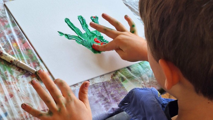 Ein kleines Kind macht einen grünen Handabdruck auf ein Blatt Papier, während es auf einer gemusterten Oberfläche sitzt.