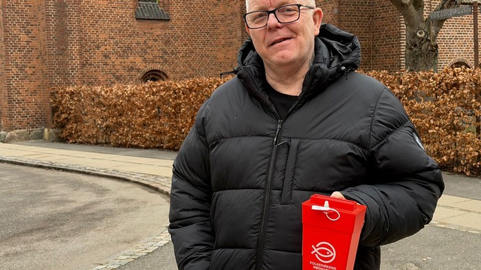 En person i en sort dunjakke holder en rød indsamlingsbøsse uden for en murstensbygning.