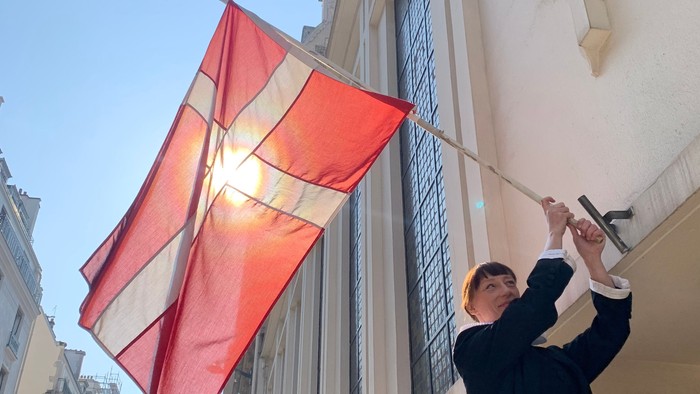 Person hejser et stort dansk flag uden for en moderne bygning på en solskinsdag.