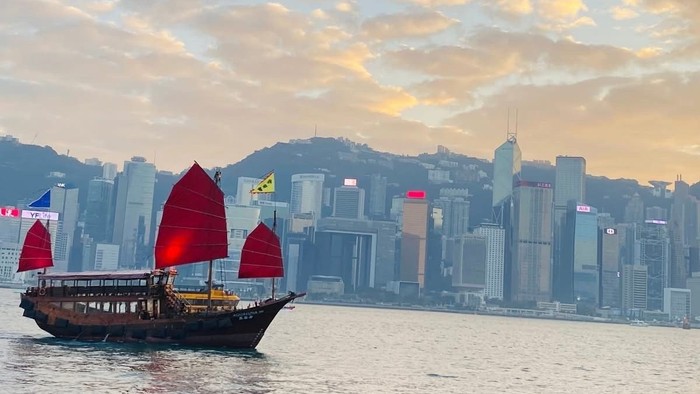 En traditionel junkbåd med røde sejl sejler forbi en moderne byskyline ved solnedgang.