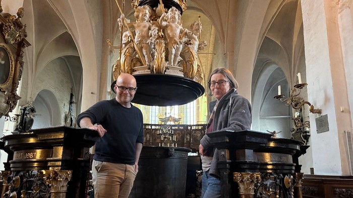 Schauspieldirektor Malte C. Lachmann steht links und Pastorin Nicola Nehmzow rechts im Bild. Beide sind am Taufbecken in St. Aegidien mit vielen Engeln über ihren Köpfen.