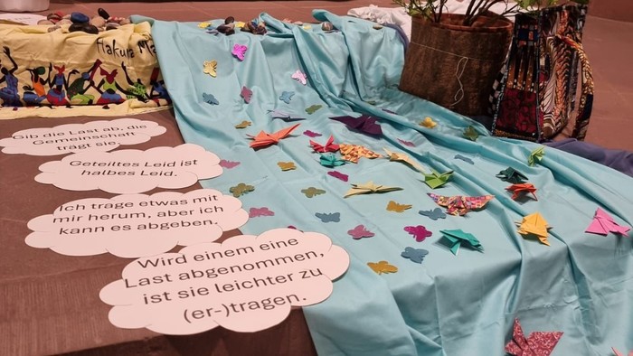 Gestaltete Gebetsecke zum Weltgebetstag mit bunten Tüchern, Origami‑Vögeln, Pflanzen und Textwolken zum Thema „Last abgeben“.