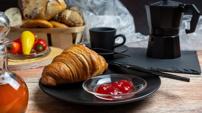 Genussvolles Frühstück mit einem Croissant mit Marmelade, Kaffee, frischem Brot und farbenfrohem Obst auf einem rustikalen Tisch.