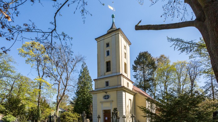 Kirchturm mit Uhr und Kreuz, umgeben von Bäumen und Friedhof