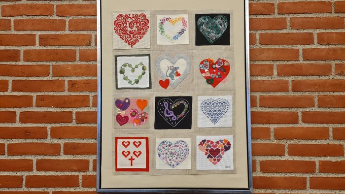 Et indrammet samling af farverige hjerteformede broderier og tekstilkunst på en murstensbaggrund.