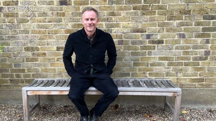 Mand i sort klædning sidder på bænk foran murstenge væg