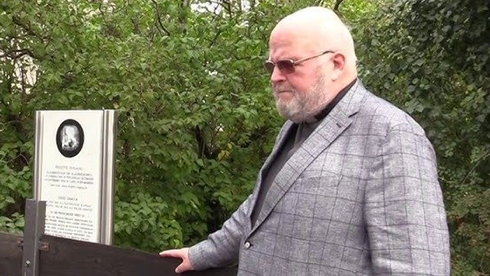 Ein glatzköpfiger Mann im Anzug zeigt auf eine Gedenktafel im Freien.