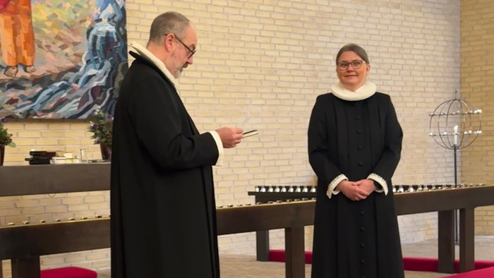 To gejstlige står i en kirke, den ene holder papirer, mens den anden iagttager ved siden af et klokkearrangement.
