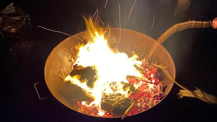Eine Person rührt nachts in einer brutzelnden Pfanne mit Essen über offener Flamme.