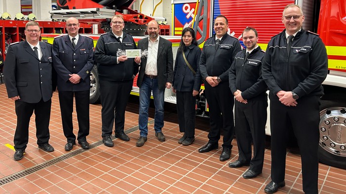 Gruppe von Feuerwehrleuten und Beamten, die sich drinnen mit einem Feuerwehrauto und einer symbolischen Scheckübergabe fotografieren lassen.