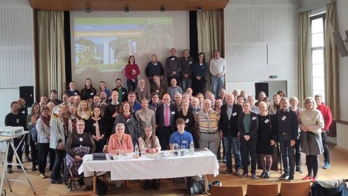 Eine große Gruppe von Menschen posiert gemeinsam für ein Gruppenfoto in einem hellen Veranstaltungssaal vor einer Bühne.