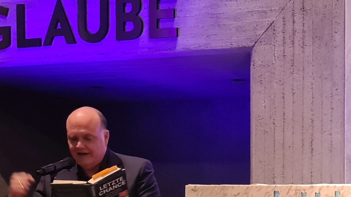 Zwei Männer sitzen auf der Bühne während einer religiösen Diskussion, mit einem Kreuz und einem Altaraufbau im Hintergrund.