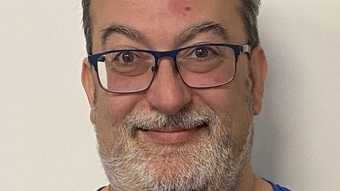 En mand med briller og skæg, der smiler og har et rødt prikke på puden.