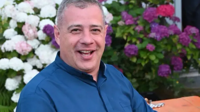 Ein lächelnder Mann in einem blauen Hemd posiert neben üppigen blühenden Sträuchern im Freien.