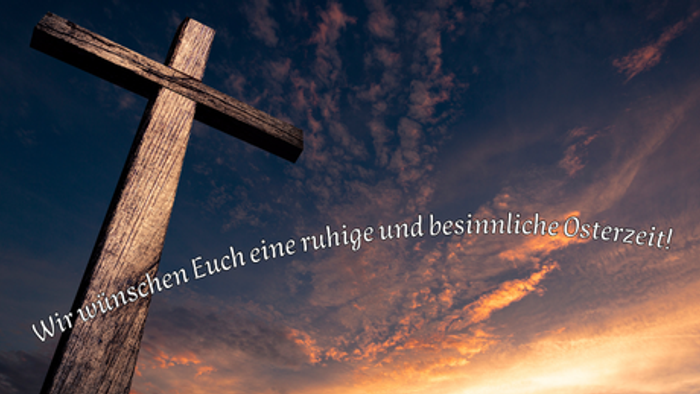 Ein hölzernes Kreuz, das sich vor einem dramatischen Sonnenuntergangshimmel abzeichnet, mit einem friedvollen Osterwunsch auf Deutsch.
