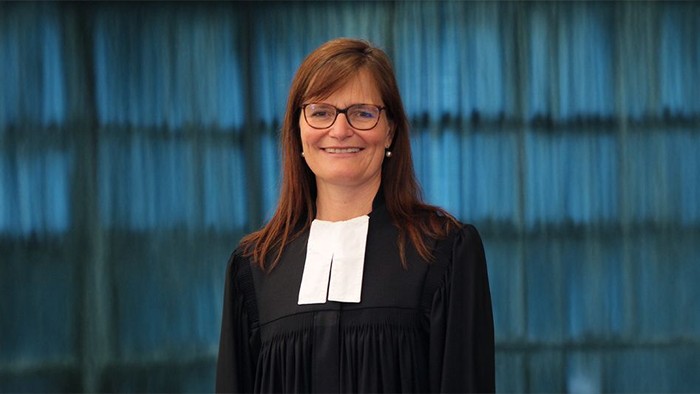 Das Bild zeigt eine Frau in schwarzem Talar mit weißem Beffchen, der typischen Amtstracht evangelischer Geistlicher. Sie steht frontal zur Kamera, lächelt freundlich und trägt eine Brille. Ihr schulterlanges Haar ist offen getragen. Der Hintergrund ist w