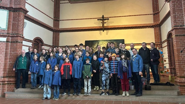 Eine große Gruppe von Kindern, Jugendlichen und Erwachsenen steht gemeinsam vor dem Altar einer Kirche mit buntem Rosettenfenster.