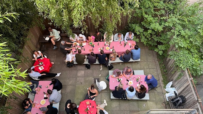 En gruppe af mennesker sidder udendørs ved røde bord i en trækirkegård.