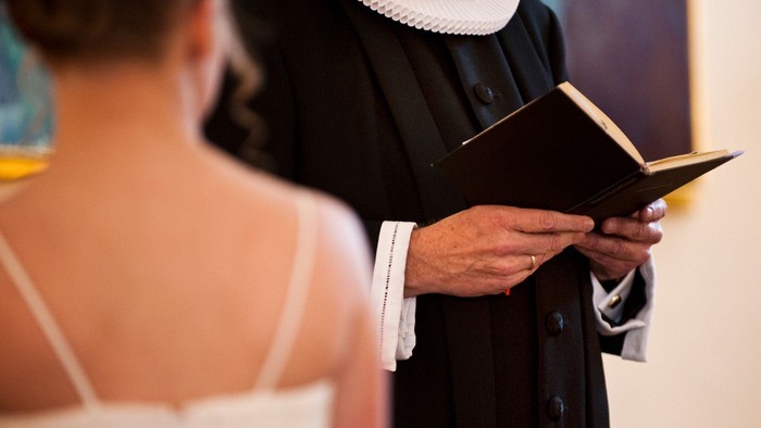 En præst i sort kappe læser fra en bog ved siden af en gift par.