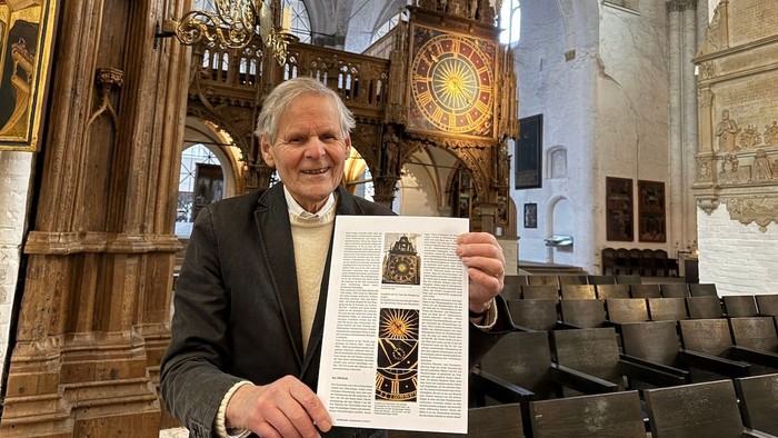 Andreas Groth steht im Dom vor dem Letten mit der Monduhr und hält eine Zeitschrift in der Hand, in der ein Artikel über die Uhr erschienen ist.