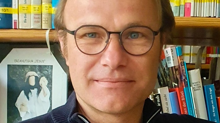 Ein Mann mit Brille und einem hohen blonden Scheitel vor einem Bücherregal