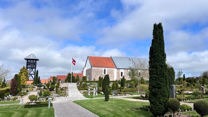 En fredfyldt kirkegård med en historisk kirke, en flagstang og velplejede grave under en blå himmel.
