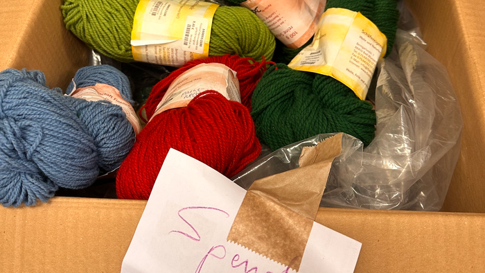 Eine Schachtel gefüllt mit verschiedenen bunten Garnen und einem handgeschriebenen Zettel mit der Aufschrift „Spindel“.