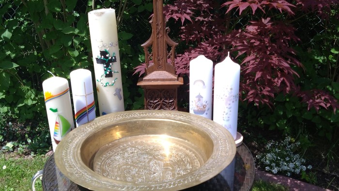 Außere Weihwasserbecken mit Kerzen, Kreuz und umgebender Begrünung.