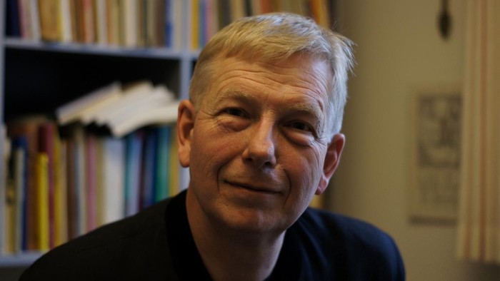 En smilende mand med gråt hår står foran en farverig bogreol og kontorindretning.