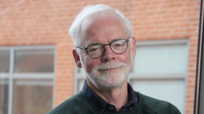 En mand med briller og gråt hår står selvsikkert med armene over kors foran murstensvægge.