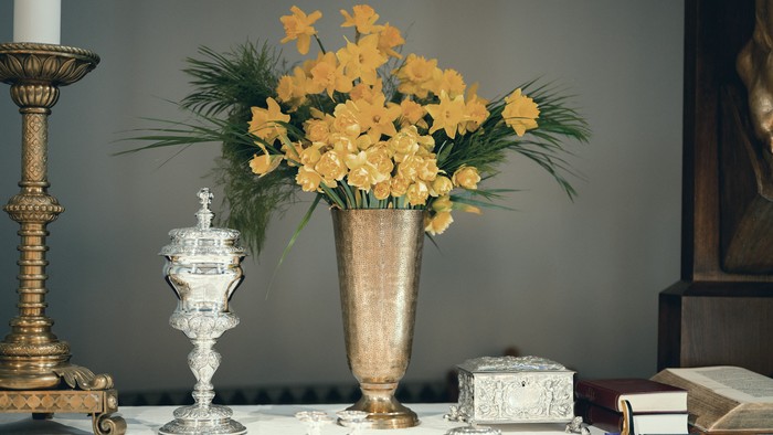 Elegant alteropsætning med en vase med gule blomster, sølvtøj og religiøse bøger på et bord.