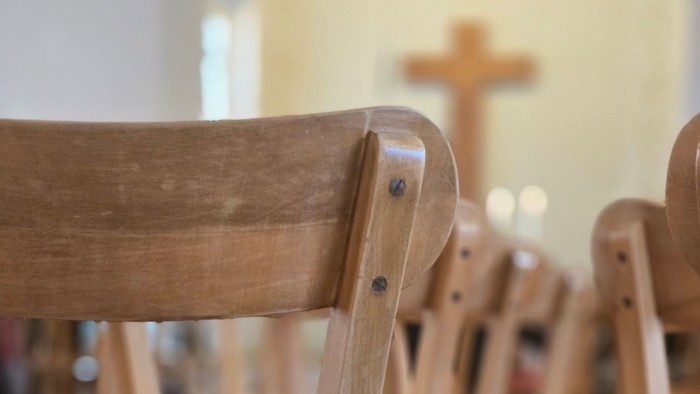 Rückansicht von hölzernen Kirchenbänken, die in ordentlichen Reihen angeordnet sind, mit einem Kreuz im Hintergrund.