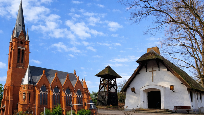 Zwei unterschiedliche Kirchengebäude – eine große neugotische Backsteinkirche und eine kleine weiß verputzte Reetdachkapelle mit Glockenturm – stehen unter einem teils wolkigen blauen Himmel nebeneinander.