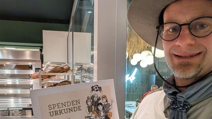 Ein Mann hält eine Spendenurkunde in einer Bäckerei mit verschiedenen Backwaren im Hintergrund.